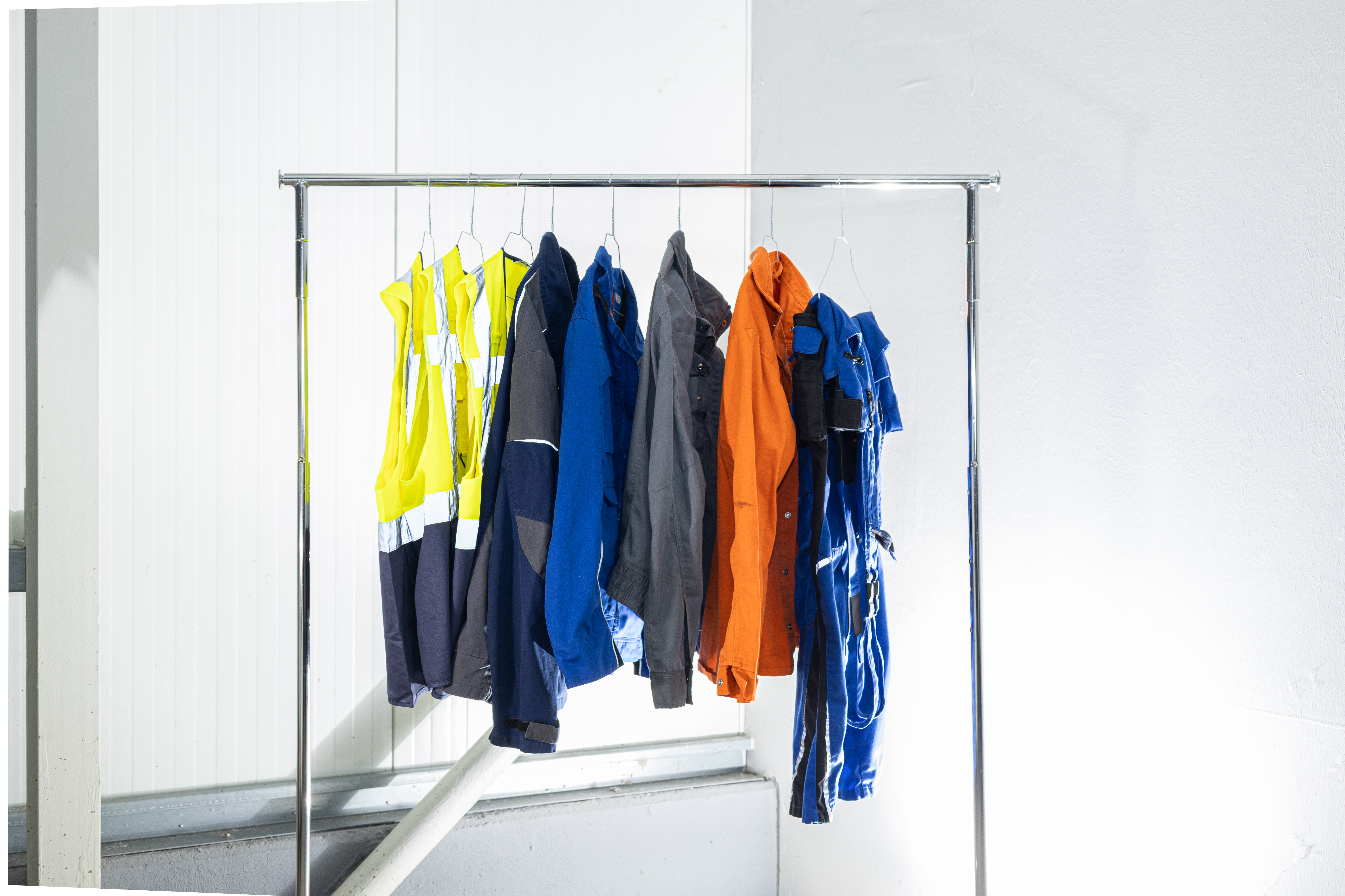 Berufs- und Schutzkleidung für Industrie und Gewerbe in Gelb, Blau, Grau und Orange hängt auf Kleiderbügeln an einer Metallstange.