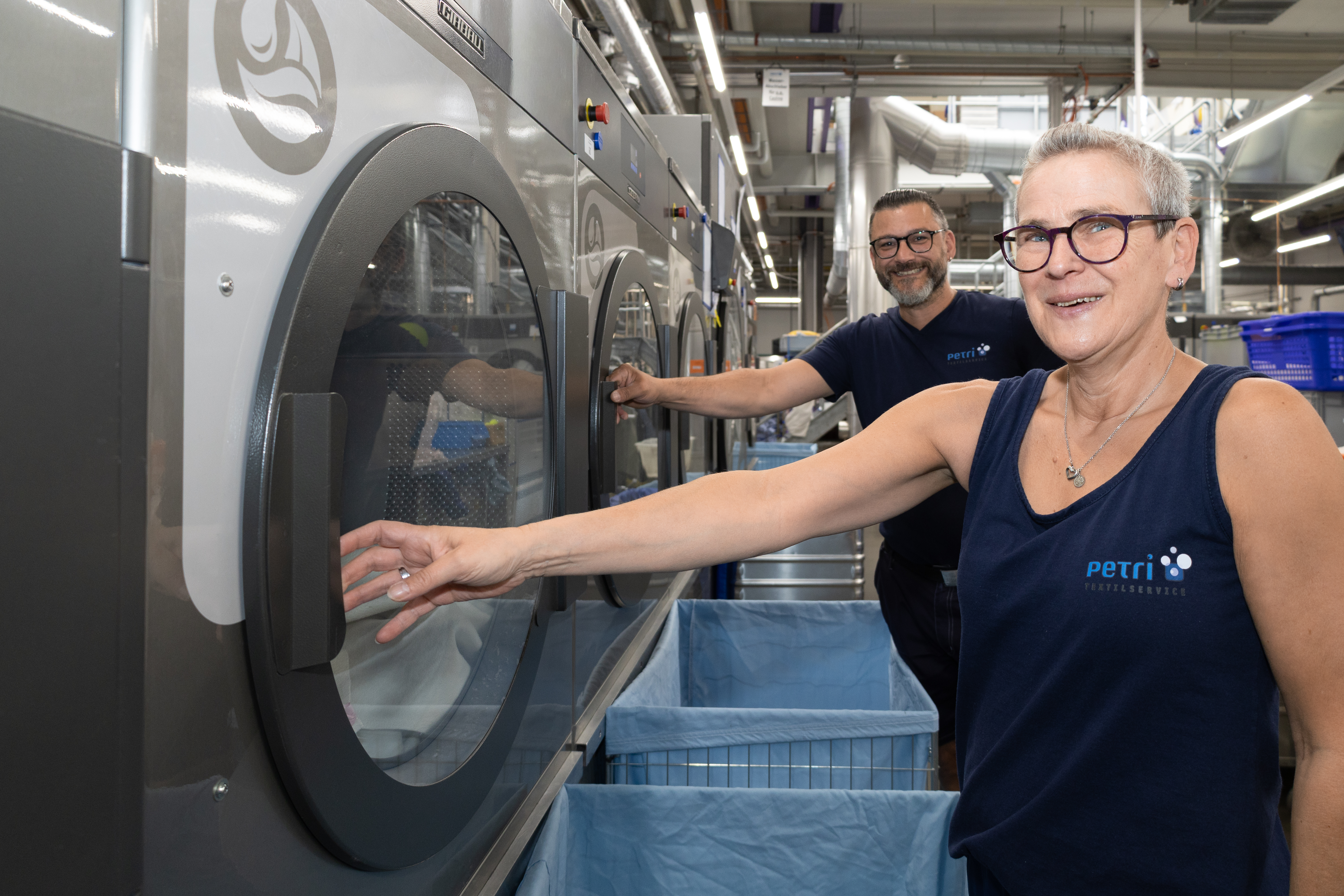 Zwei Mitarbeitende von Textilservice Petri arbeiten an modernen Industriewaschmaschinen in der Wäscherei.