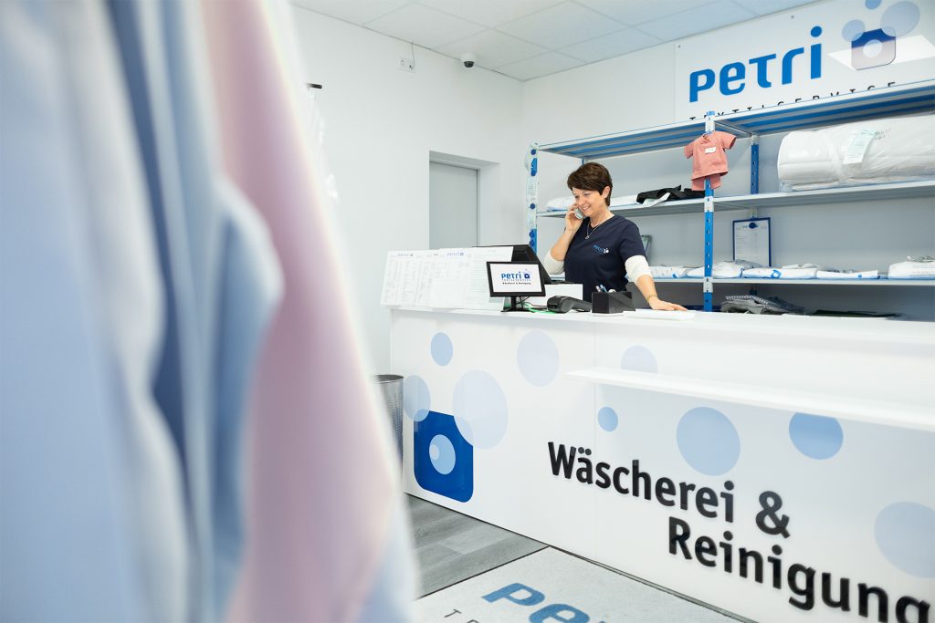 Lächelnde Mitarbeiterin in dunkelblauem Petri-Shirt telefoniert am weißen Empfangstresen einer Petri Textilservice Annahmestelle mit der Aufschrift „Wäscherei & Reinigung“. Im Hintergrund stehen blaue Regale mit verpackter Wäsche, im Vordergrund hängen unscharf Textilien in Blau und Rosa.